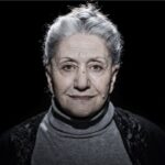 A grayscale portrait of an elderly woman with short gray hair, wearing a high-neck sweater and a black shawl. She stands against a dark background with a hopeful expression. Text below reads: "Suraja / Age: 69 / Azerbaijan. I wish for a good life in the Netherlands.