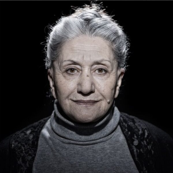 A grayscale portrait of an elderly woman with short gray hair, wearing a high-neck sweater and a black shawl. She stands against a dark background with a hopeful expression. Text below reads: "Suraja / Age: 69 / Azerbaijan. I wish for a good life in the Netherlands.