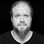 A bearded man with a receding hairline looks slightly upwards with a concerned or perplexed expression. He is wearing a black shirt and is set against a black background. The image is in black and white.