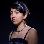A young woman stands against a dark background, looking slightly to the side. She has dark hair tied back with a headband, wears a sleeveless patterned dress, and a beaded necklace. She has a calm, composed expression with a touch of red lipstick.