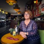 A woman with glasses is sitting at a table in a cozy pub. She is smiling and wearing a blue blouse with white polka dots. The pub is warmly lit, with various posters and decorations on the red and brown walls. Other tables and a bar are visible in the background.