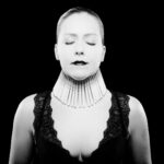 A black-and-white photo of a person with closed eyes and dark lipstick, wearing an intricate neckpiece and a black lace top. The neckpiece consists of many vertical rods, creating a structured, collar-like appearance. The background is dark, highlighting the subject.