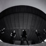 A black-and-white image showing five people standing spaced out on a curved platform in front of a large, dome-shaped structure that looks like a UFO. The person in the center strikes a confident pose, while the others stand casually, reflecting an enigmatic and bold ambiance.