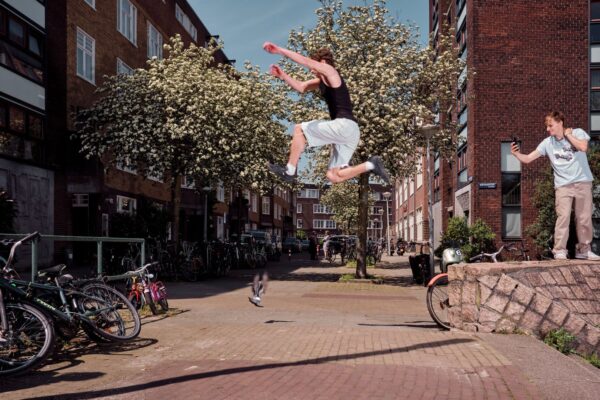A person captured mid-air while performing a parkour jump between buildings in an urban setting. Another person on a low wall enthusiastically watches the jump. Bicycles are parked along the sidewalk, and there are flowering trees lining the street.