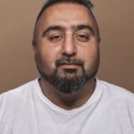 A middle-aged man with short dark hair, a trimmed beard, and mustache poses against a plain brown background while wearing a white t-shirt. He is facing the camera with a neutral expression.