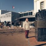 An elderly man stands beside a black marble monument with a Star of David, in a busy square in front of a modern building with “Almada” signage, as people walk by.