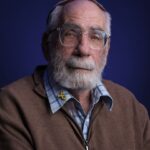 An elderly man with a white beard and glasses wears a brown sweater over a blue plaid shirt, featuring a yellow ribbon pin. He has a neutral expression and sits against a dark blue background.