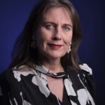 A woman with long brown hair, wearing floral earrings and a black and white patterned blouse, poses against a dark blue background. She has light eyes and red lipstick, looking directly at the camera with a neutral expression.