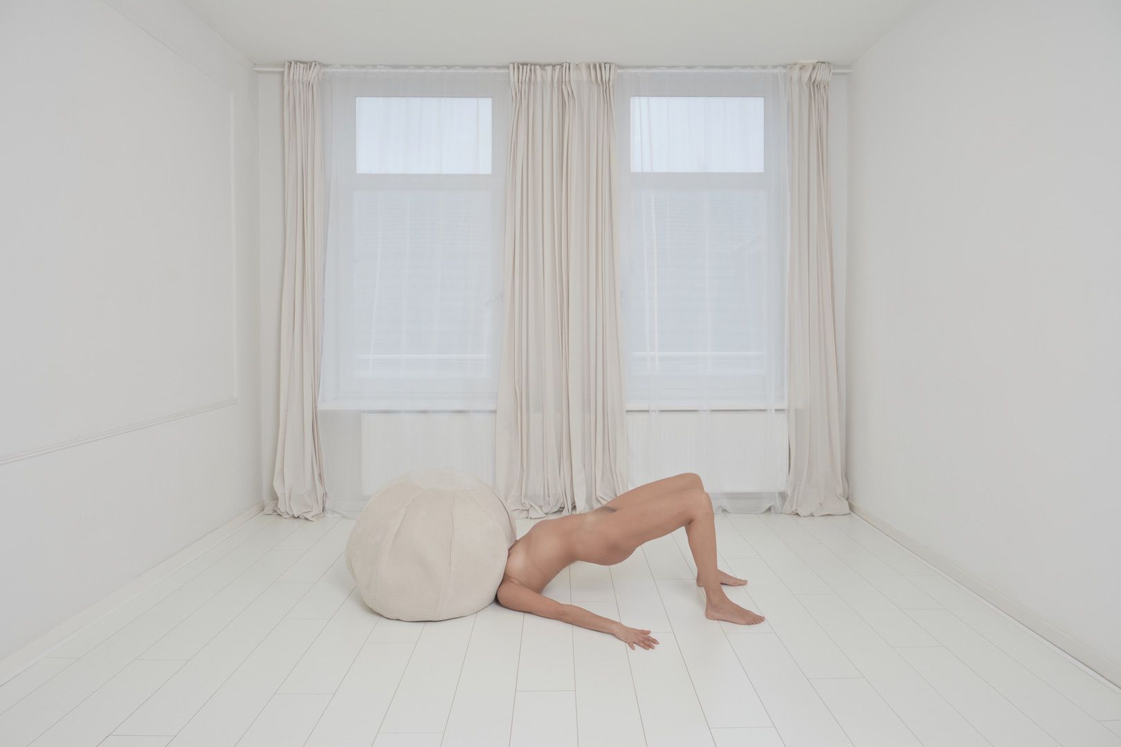 A nude person lies on their back on a white floor, head obscured by a large, round, beige cushion in a minimalistic white room with tall windows and sheer curtains—a striking addition to any artistic portfolio.