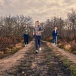 Three people jogging outdoors on a dirt path through a wooded area with leafless trees, under a cloudy sky. They appear to be dressed in athletic clothing, enjoying a run together in nature.