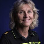 A smiling woman with shoulder-length blonde hair wears a black and yellow police uniform, standing against a dark blue background.
