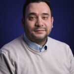 A man with short dark hair and a trimmed beard wearing a light beige sweater over a light blue collared shirt, posing in front of a dark blue background.
