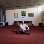 A man sits cross-legged on a red carpet in a spacious room with framed portraits on the wall, cushions along the walls, and sunlight streaming through high windows.