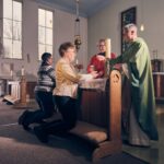 Two people kneel at an altar rail in a church, receiving communion from two clergy members; sunlight streams through large windows and stained glass in the background.