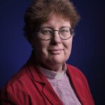 A person with short, curly brown hair and glasses, wearing a red jacket and a clerical collar, poses against a dark blue background, looking directly at the camera with a slight smile.
