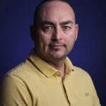 A man with short dark hair and light skin wearing a yellow polo shirt poses against a dark blue background, looking directly at the camera with a neutral expression.