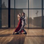 A woman in a red dress and a man in a gray suit dance together in a sunlit room with large windows, wooden floor, and a potted plant on the left.