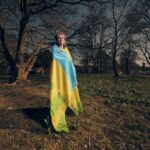 An older person stands outdoors in a leafless park, wrapped in a large blue and yellow flag, with sunlight casting long shadows on the ground.