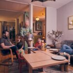 A family gathers in a cozy living room. Two adults and two children sit around a coffee table as an older man, standing, serves popcorn from a large bowl. The room is warmly lit and decorated with art and plants.