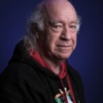 An older man with thinning gray hair and a receding hairline, wearing a black hoodie with a colorful graphic, poses against a dark blue background. He has a neutral expression and is looking slightly toward the camera.