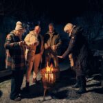 Four adults in winter coats gather outside around a fire pit at night, holding drinks and warming themselves. A warmly lit building is behind them, and bare trees are visible in the background.