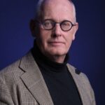 A bald older man with round blue glasses wears a black turtleneck and a checked blazer, posing against a dark blue background.
