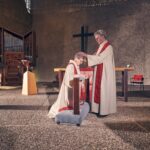 A priest in white and red robes kneels at an altar rail while another priest stands, laying hands on their head in blessing inside a modern church with a cross, organ, and rainbow banner in the background.