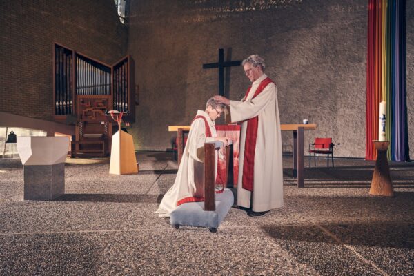 A priest in white and red robes kneels at an altar rail while another priest stands, laying hands on their head in blessing inside a modern church with a cross, organ, and rainbow banner in the background.