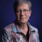 An older woman with short gray hair and glasses, wearing a colorful floral-patterned jacket and a single dangling earring, sits against a dark blue background, looking calmly at the camera.