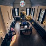 A person sits alone on a bench inside a spacious, high-ceilinged synagogue with tall windows, dark wooden pews, chandeliers, and a colorful stained glass window overhead.