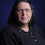 A middle-aged woman with shoulder-length dark hair, glasses, and a neutral expression poses against a dark blue background. She is wearing a black cardigan over a patterned blouse.
