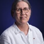 A person with gray hair tied back, wearing round glasses and a white button-up shirt, smiles gently against a dark blue background.