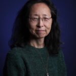 A woman with long dark hair and glasses wearing a green sweater and a pendant necklace, poses in front of a dark blue background.