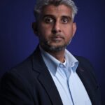 A man with short gray hair and a beard is wearing a dark blazer over a light blue shirt, standing against a dark blue background, looking directly at the camera with a serious expression.