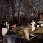 A person dressed in white stands among gravestones in a sunlit cemetery, holding flowers. Leafless trees and shrubs surround the area, casting shadows on the ground.