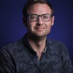 A man with short brown hair and glasses wears a patterned dark shirt and looks at the camera with a slight smile, posed against a solid dark blue background.