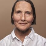 A middle-aged woman with short brown hair and blue eyes wears a white collared shirt, looking directly at the camera with a calm expression against a plain beige background.