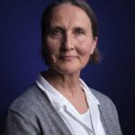 Portrait of an older woman with short brown hair wearing a white shirt and gray cardigan, smiling softly against a dark blue background.