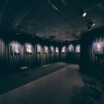 A dimly lit gallery room displays framed portraits of various people on black curtains, with spotlights illuminating each photo. A few empty chairs are arranged in the foreground.