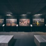 A dimly lit art gallery displays five large photographs on black curtains, each showing different indoor or outdoor scenes. White benches and information stands are placed in front of the artworks.