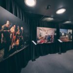 Three large photographs are displayed on a black curtain wall in a dimly lit gallery space, each mounted above a stand with information placards. The room has spotlights and a carpeted floor.