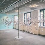 An art installation in a bright room features hundreds of white paper cranes suspended in midair between two metal poles, creating the illusion of a flock flying through the space. Large windows and a blue wall are visible in the background.