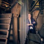 A woman in a purple jacket stands with arms crossed inside a wooden clock tower, surrounded by large gears, cables, and wooden stairs. Warm light filters through the machinery.
