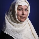 An older woman wearing a white hijab and dark clothing poses in front of a deep blue background, looking calmly at the camera with a gentle expression.