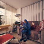 A woman wearing a blue hijab and patterned pants sits on a beige sofa, reading a book in a cozy living room with warm lighting, cushions, houseplants, and a window letting in daylight.