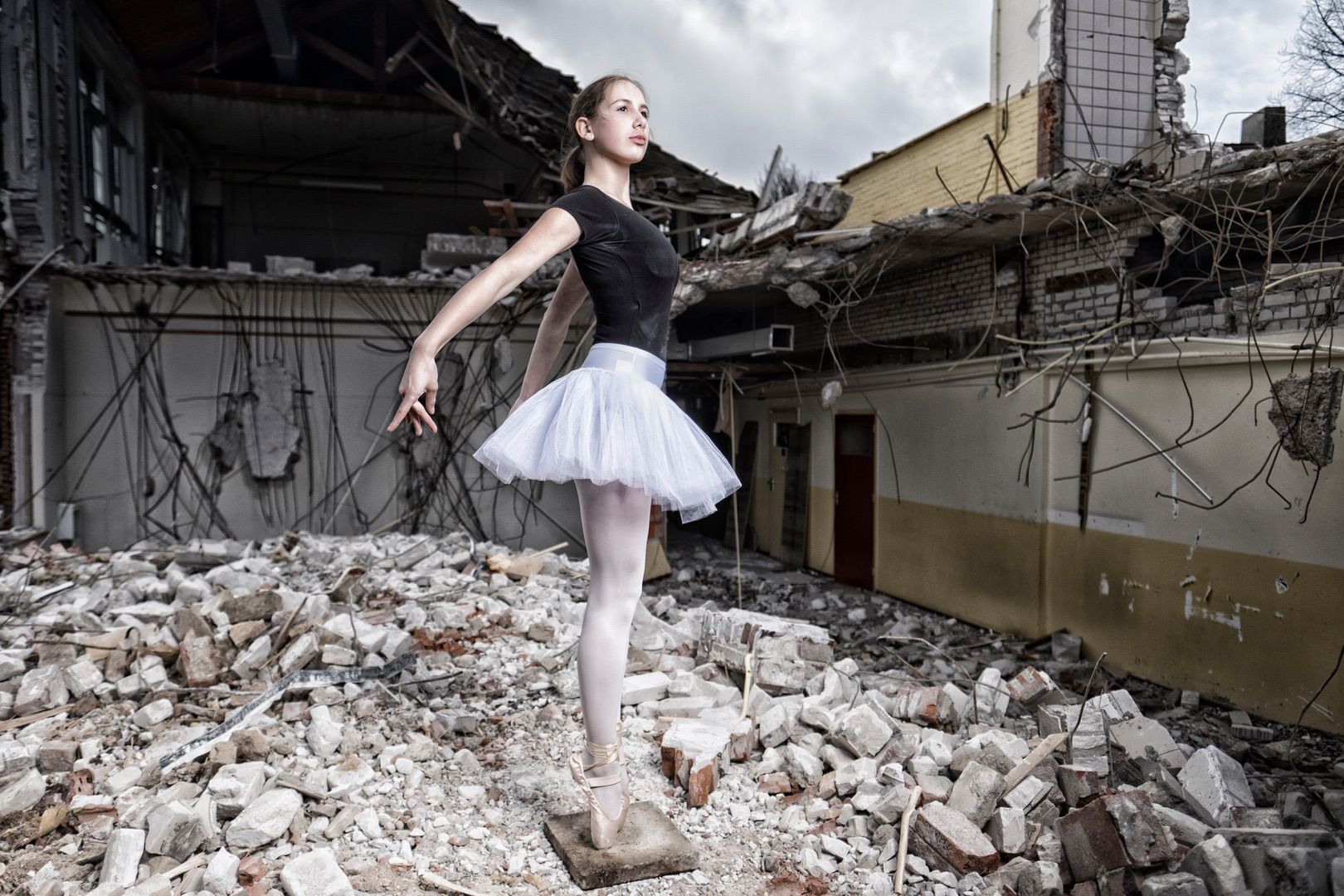 A young ballerina in a white tutu and black leotard poses gracefully on pointe amid rubble, creating a striking portfolio image that contrasts beauty and devastation in a destroyed building.