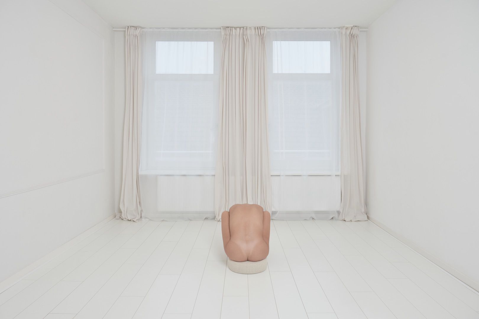 A minimalist white room with large windows and sheer curtains; a nude, headless mannequin torso kneels on the light wooden floor, creating a striking centerpiece—perfect for a portfolio highlighting clean, modern design.
