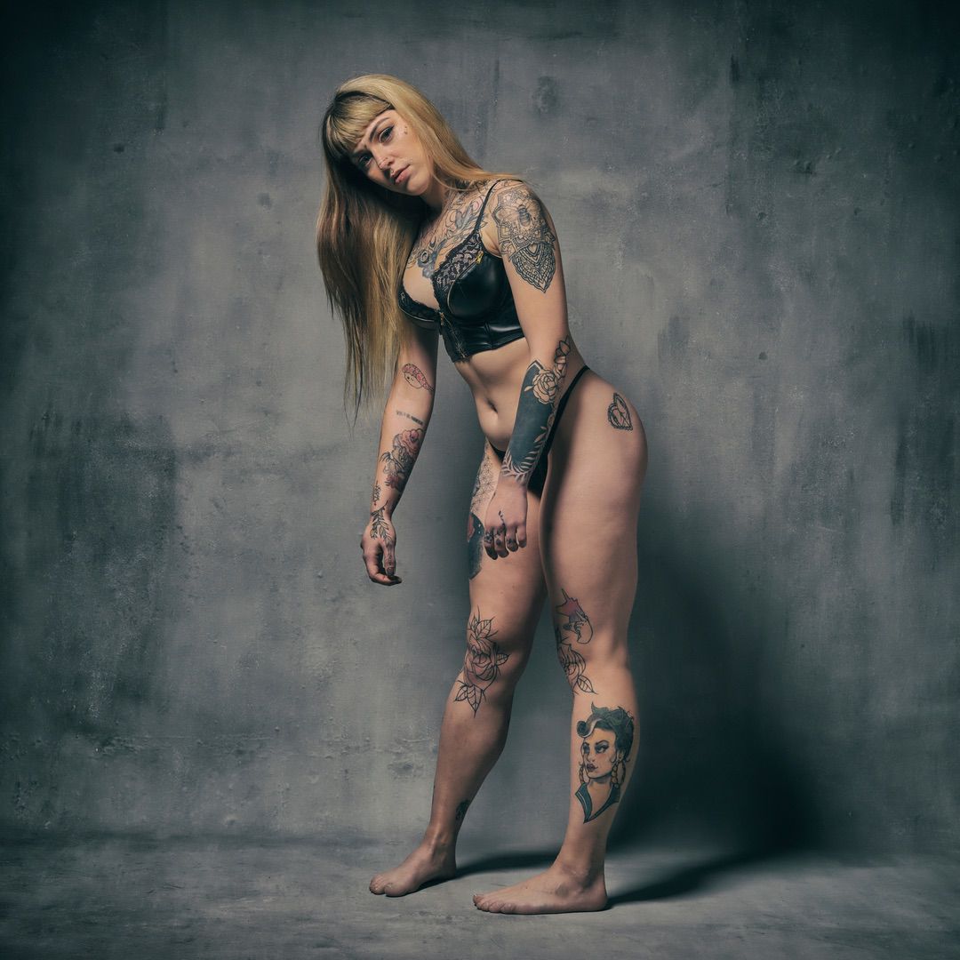 A woman with long blonde hair and many tattoos poses barefoot in black lingerie against a textured gray background, slightly leaning and looking at the camera—a striking image perfect for a creative portfolio.