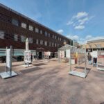 A spacious outdoor plaza with art displays in glass cases, people walking, and a modern building with horizontal slats in the background under a blue sky with scattered clouds.