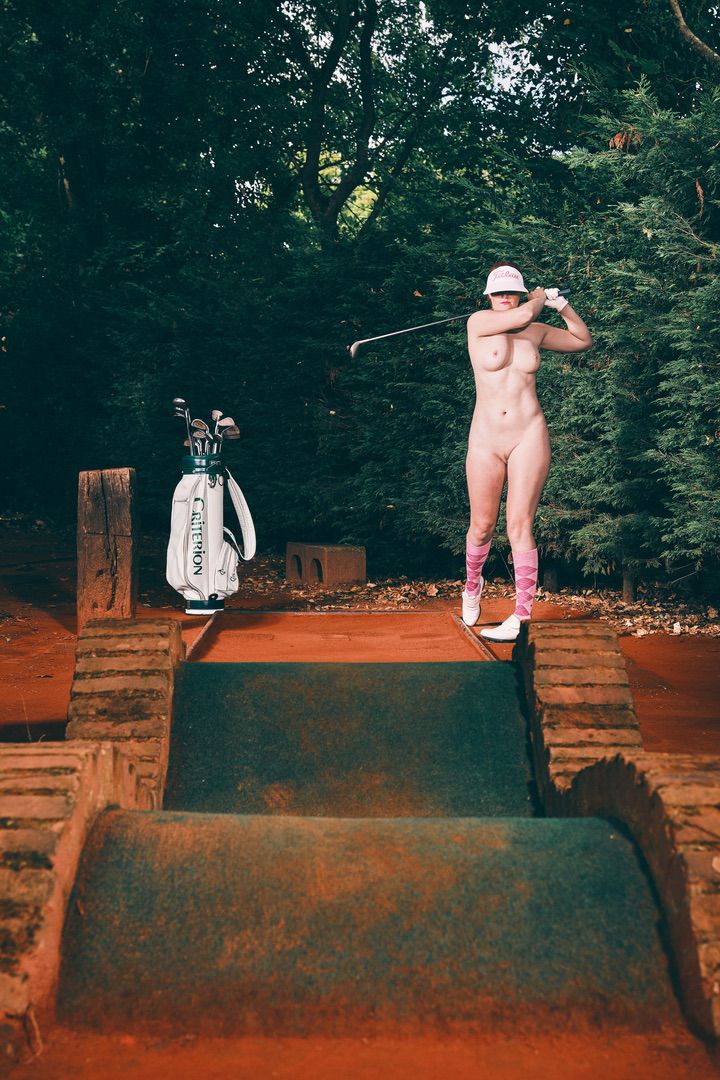 A portfolio-worthy shot: a person wearing only a cap, pink socks, and shoes swings a golf club on a small outdoor platform, with a golf bag and dense trees forming the backdrop.